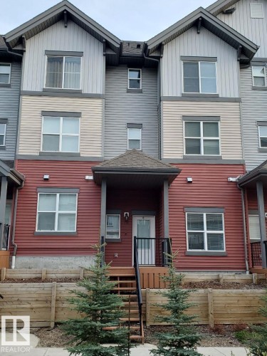Multi-level townhome featuring a covered entry, horizontal siding in varying shades, dark framed windows, and a tiered wooden retaining wall with planted evergreens - 118 655 Watt Boulevard, Edmonton, AB - Outdoor