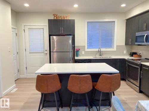 Kitchen featuring a central island with a white countertop, dark wood-finish cabinetry, stainless steel appliances, light wood-finish flooring, and a tiled backsplash - 118 655 Watt Boulevard, Edmonton, AB - Indoor Photo Showing Kitchen