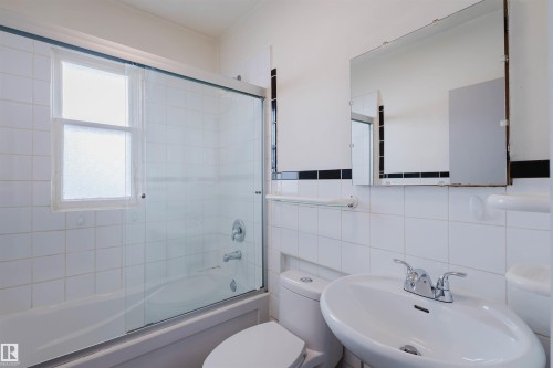 Bathroom featuring a white ceramic tile surround, a single-pane window with frosted glass, a chrome faucet and showerhead, a sliding glass shower door, and a wall-mounted rectangular mirror - 10511 80 Street, Edmonton, AB - Indoor Photo Showing Bathroom