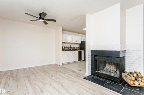 Living area featuring wood-finish flooring, a contemporary ceiling fan with integrated lighting, and a white painted brick fireplace with a black hearth - 9221 172 Street, Edmonton, AB - Indoor Photo Showing Living Room With Fireplace