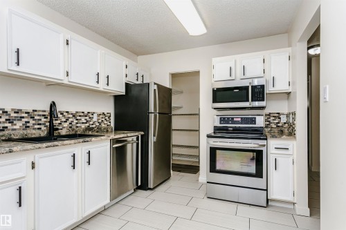 Kitchen featuring white cabinetry with black hardware, mosaic tile backsplash, black sink with matte black faucet, stainless steel appliances, and light-toned tile flooring - 9221 172 Street, Edmonton, AB - Indoor Photo Showing Kitchen With Stainless Steel Kitchen