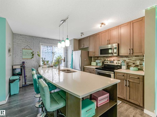 Modern kitchen featuring a large curved island with an undermount double sink, stainless steel appliances, wood-finish cabinetry, and a mosaic tile backsplash - 44 1140 Chappelle Boulevard, Edmonton, AB - Indoor Photo Showing Kitchen With Stainless Steel Kitchen With Double Sink