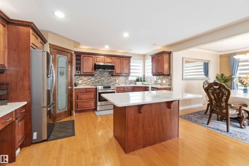 Kitchen featuring wood-finish cabinetry, stainless steel appliances, a center island with a light-toned countertop, a tile backsplash, and light wood-finish flooring - 6116 53 Avenue, Beaumont, AB - Indoor Photo Showing Kitchen With Stainless Steel Kitchen With Upgraded Kitchen