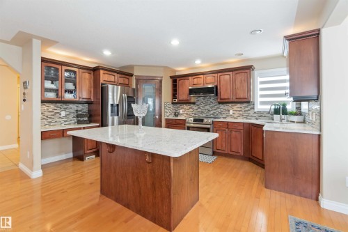 Kitchen featuring a central island with a light-toned countertop, wood-finish flooring, warm wood cabinetry, and a mosaic tile backsplash - 6116 53 Avenue, Beaumont, AB - Indoor Photo Showing Kitchen With Upgraded Kitchen
