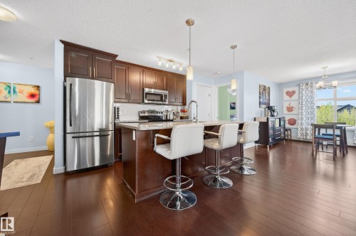 Open-concept kitchen featuring wood-finish flooring, dark wood cabinetry, stainless steel appliances, and an eat-at island with pendant lighting - 340 6076 Schonsee Way, Edmonton, AB - Indoor Photo Showing Kitchen With Stainless Steel Kitchen With Upgraded Kitchen