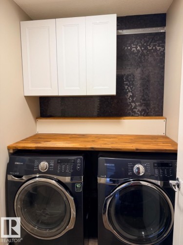 Laundry area featuring a wood-finish countertop, three upper white shaker-style cabinets, and a dark hexagonal tile backsplash - 648 178 Street, Edmonton, AB - Indoor Photo Showing Laundry Room