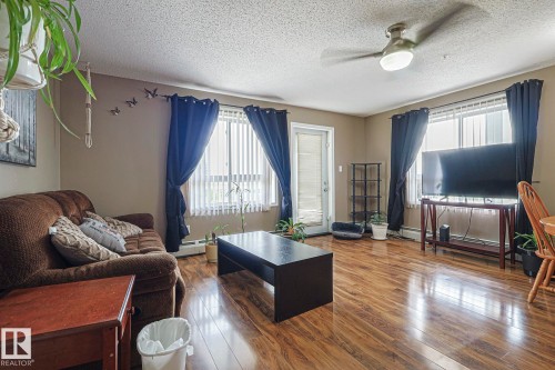 Spacious living area featuring wood-finish flooring, a ceiling fan with integrated lighting, and multiple windows with vertical blinds - 314 5340 199 Street, Edmonton, AB - Indoor Photo Showing Living Room