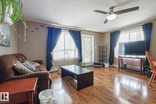Living area featuring wood-finish flooring, a ceiling fan with integrated lighting, and multiple windows with vertical blinds - 314 5340 199 Street, Edmonton, AB - Indoor Photo Showing Living Room