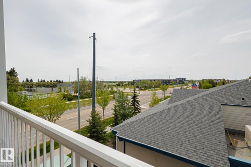 Balcony railing offering an elevated perspective of the surrounding area, with a paved road, green spaces, and distant buildings under an open sky - 314 5340 199 Street, Edmonton, AB - Outdoor With View
