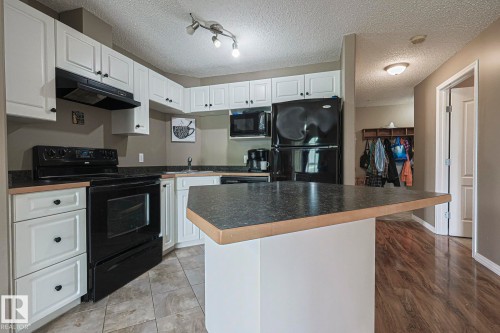 Kitchen featuring white cabinetry with black hardware, black appliances, a central island with an extended countertop, and tile flooring - 314 5340 199 Street, Edmonton, AB - Indoor Photo Showing Kitchen