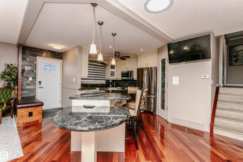 This open-concept area features polished hardwood flooring, a kitchen island with a stone countertop, pendant lighting, and stainless steel appliances - 9831 185 Street, Edmonton, AB - Indoor Photo Showing Kitchen With Upgraded Kitchen