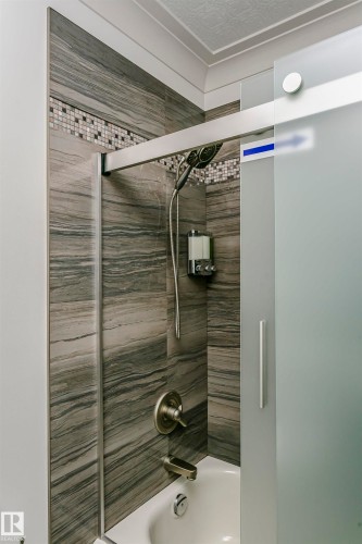 Bathroom featuring a bathtub with a shower, dark gray tiled walls with a mosaic accent, and a frosted glass shower door - 9831 185 Street, Edmonton, AB - Indoor Photo Showing Bathroom