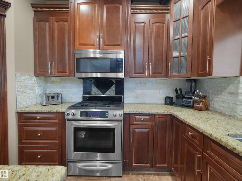 Kitchen featuring stainless steel appliances, wood-finish cabinetry, speckled countertops, and a stacked stone backsplash - 1511 69 Street, Edmonton, AB - Indoor Photo Showing Kitchen