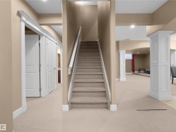 Carpeted staircase with white railings, leading to an expansive lower level featuring cream-colored carpeting, white trim, recessed lighting, and decorative white columns - 