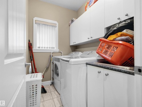 Dedicated laundry area featuring tile flooring, a window with vertical blinds, upper and lower white cabinetry with silver hardware, and a built-in sink with a dark countertop - 1511 69 Street, Edmonton, AB - Indoor Photo Showing Laundry Room