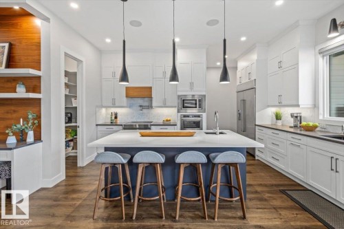 Gourmet kitchen featuring a large central island with seating, white shaker-style cabinetry, stainless steel appliances, wood-finish flooring, and a walk-in pantry - 10611 146 Street Nw, Edmonton, AB - Indoor Photo Showing Kitchen With Upgraded Kitchen