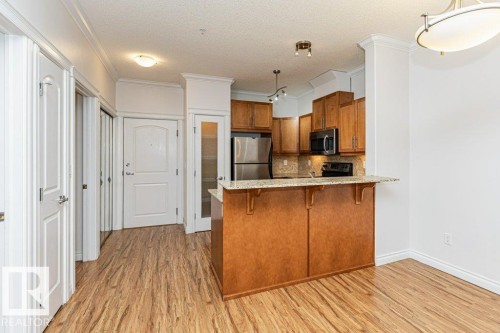 Open-concept kitchen featuring wood cabinetry, stainless steel appliances, a stone countertop island with breakfast bar, and wood-finish flooring - 145 10121 80 Avenue, Edmonton, AB - Indoor Photo Showing Kitchen