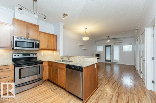 Kitchen featuring wood-finish flooring, stainless steel appliances, light wood cabinetry, and a granite-style countertop with an integrated double basin sink - 145 10121 80 Avenue, Edmonton, AB - Indoor Photo Showing Kitchen With Stainless Steel Kitchen With Double Sink