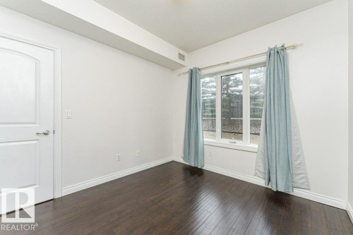 Spacious room featuring dark wood-finish flooring, a large multi-pane window, bright white walls, and elegant white baseboards - 145 10121 80 Avenue, Edmonton, AB - Indoor Photo Showing Other Room