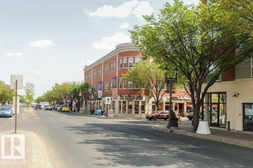 Streetscape featuring a red brick building with a rounded corner, street-level retail spaces, pedestrian-friendly sidewalks, mature street trees, and a multi-lane roadway - 145 10121 80 Avenue, Edmonton, AB - Outdoor