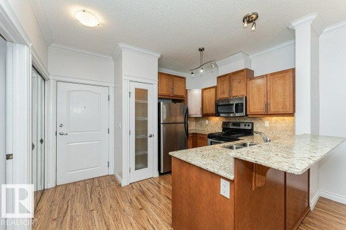 Kitchen featuring stainless steel appliances, wood-finish cabinetry, a dual basin sink, light granite countertops, and wood-finish flooring - 145 10121 80 Avenue, Edmonton, AB - Indoor Photo Showing Kitchen With Stainless Steel Kitchen