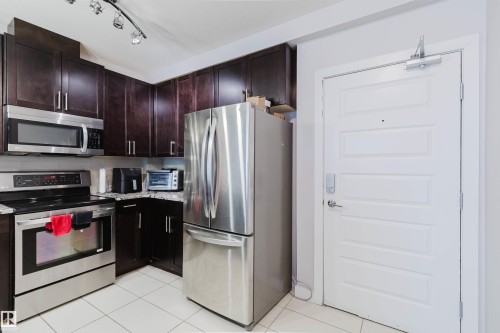 Kitchen featuring dark wood-finish cabinetry, stainless steel appliances, granite-style countertops, and white tile flooring - 510 5151 Windermere Boulevard, Edmonton, AB - Indoor Photo Showing Kitchen With Stainless Steel Kitchen