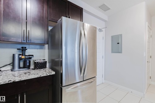 Kitchen with dark wood-finish cabinetry, stainless steel refrigerator, stone countertop, and light-toned tile backsplash - 510 5151 Windermere Boulevard, Edmonton, AB - Indoor Photo Showing Kitchen