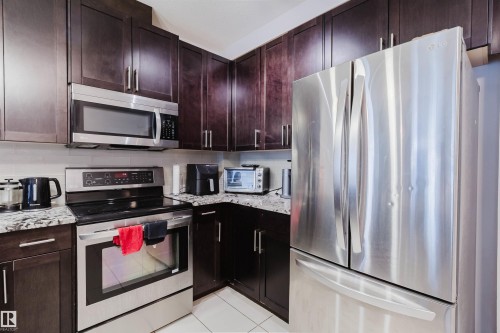 Modern kitchen featuring dark wood cabinetry, stainless steel appliances, granite-style countertops, and a subway tile backsplash - 510 5151 Windermere Boulevard, Edmonton, AB - Indoor Photo Showing Kitchen With Stainless Steel Kitchen