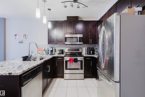 Kitchen featuring dark wood cabinetry, light-toned granite-style countertops, and stainless steel appliances including a range, microwave, and refrigerator - 510 5151 Windermere Boulevard, Edmonton, AB - Indoor Photo Showing Kitchen With Stainless Steel Kitchen With Upgraded Kitchen