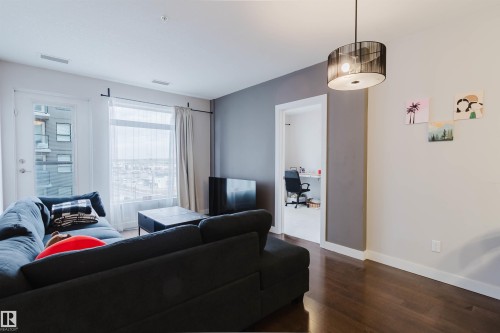 Spacious living area featuring dark wood-finish flooring and a prominent window with sheer and solid curtains - 510 5151 Windermere Boulevard, Edmonton, AB - Indoor Photo Showing Living Room