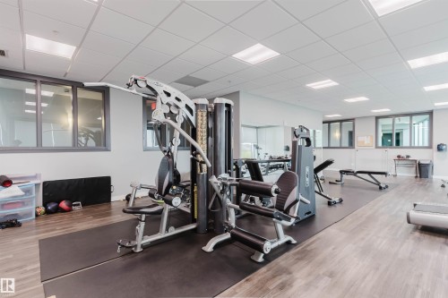 Dedicated fitness center featuring wood-finish flooring, a suspended acoustic tile ceiling with integrated lighting, multiple pane windows, a full-length wall mirror, and a central exercise machine - 510 5151 Windermere Boulevard, Edmonton, AB - Indoor Photo Showing Gym Room