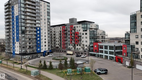Contemporary multi-story building featuring a blue and grey facade, individual balconies with glass railings, a circular drive with central landscaping, and a street-level entrance - 510 5151 Windermere Boulevard, Edmonton, AB - Outdoor With Facade