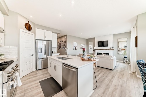 Open-concept main level featuring light wood-finish flooring, a contemporary kitchen island with a double sink, stainless steel appliances, white cabinetry, and a fireplace with a wood mantle and integrated television mount - 38 Prospect Place, Spruce Grove, AB - Indoor Photo Showing Kitchen With Double Sink With Upgraded Kitchen