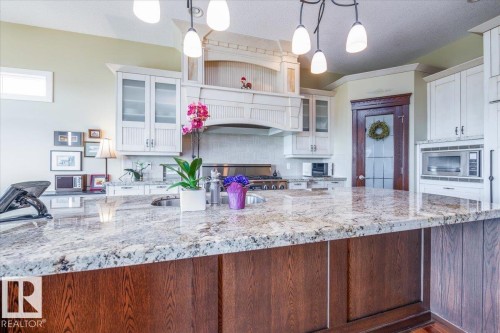 Spacious kitchen featuring a large island with a stone-finish countertop, dark wood-finish cabinetry, white upper cabinetry with glass inserts, a built-in range, and a decorative range hood - 2413 Tegler Green, Edmonton, AB - Indoor Photo Showing Kitchen With Upgraded Kitchen