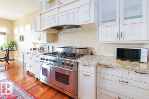 Kitchen featuring a stainless steel range with double ovens, a built-in range hood, white cabinetry with frosted glass inserts, light granite countertops, and wood-finish flooring - 2413 Tegler Green, Edmonton, AB - Indoor Photo Showing Kitchen With Upgraded Kitchen