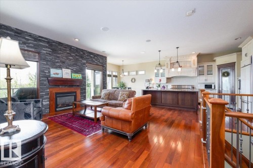 Spacious living area featuring rich wood-finish flooring, a stone-accented fireplace with a wood mantel, a kitchen island with pendant lighting, and a stairwell with wood newel posts and metal spindles - 2413 Tegler Green, Edmonton, AB - Indoor With Fireplace