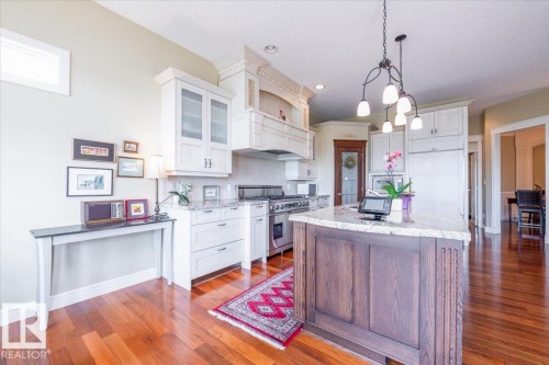 Gourmet kitchen featuring a substantial island with a light-toned countertop, a range with an elegant vent hood, wood-finish flooring, white cabinetry with glass-front uppers, and a decorative pendant light fixture - 2413 Tegler Green, Edmonton, AB - Indoor Photo Showing Kitchen