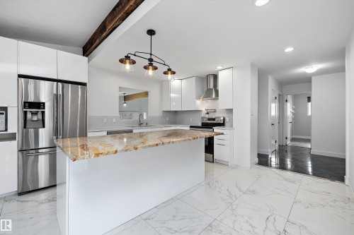 Contemporary kitchen featuring a large island with a patterned countertop, white flat-panel cabinetry, a stainless steel refrigerator, and a decorative exposed wood beam - 11525 37A Avenue Nw, Edmonton, AB - Indoor Photo Showing Kitchen With Upgraded Kitchen