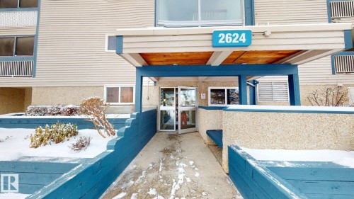 Building entrance with a teal and beige color palette, featuring a prominent address number (2624), an overhead canopy with a wood-finish underside, and an accessible ramp leading to the double glass entry doors - 402 2624 Millwoods East Road E, Edmonton, AB -  With Exterior