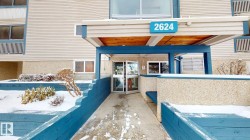 Building entrance with a teal and beige color palette, featuring a prominent address number (2624), an overhead canopy with a wood-finish underside, and an accessible ramp leading to the double glass entry doors - 
