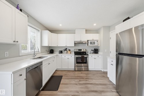 Modern kitchen featuring white shaker cabinetry, stainless steel appliances, light countertops, wood-finish flooring, and recessed lighting - 11 3380 28A Avenue Nw, Edmonton, AB - Indoor Photo Showing Kitchen With Stainless Steel Kitchen With Double Sink