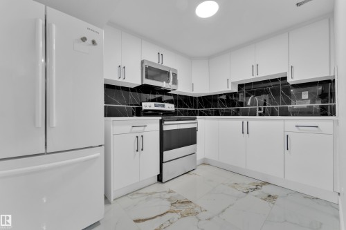 Bright kitchen featuring white cabinetry, black marble-look backsplash, stainless steel appliances, and large format floor tiling - 5118 17A Avenue Nw, Edmonton, AB - Indoor Photo Showing Kitchen