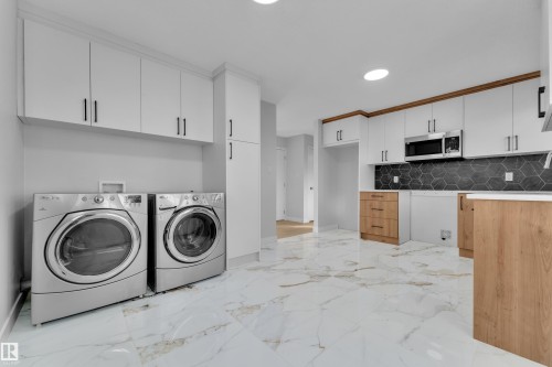 Spacious room featuring large format tile flooring, white upper cabinetry with dark hardware, a built-in microwave, and a dark hexagonal tile backsplash - 5118 17A Avenue Nw, Edmonton, AB - Indoor Photo Showing Laundry Room
