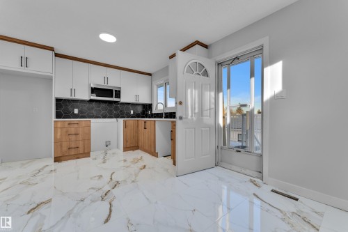 Kitchen featuring marble-look flooring, two-tone cabinetry with wood-finish lower cabinets and white upper cabinets, a dark hexagonal tile backsplash, stainless steel microwave, and a white exterior door with an arched window - 5118 17A Avenue Nw, Edmonton, AB - Indoor Photo Showing Kitchen