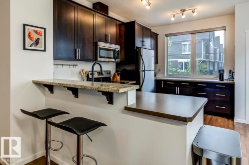 Modern kitchen featuring dark wood-finish cabinetry, stainless steel appliances, a granite breakfast bar, and wood-finish flooring - 67 603 Watt Boulevard, Edmonton, AB - Indoor Photo Showing Kitchen