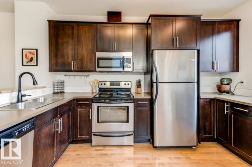 Kitchen featuring wood-finish flooring, dark wood cabinetry, light-toned countertops, a stainless steel refrigerator, and a stainless steel oven/range - 67 603 Watt Boulevard, Edmonton, AB - Indoor Photo Showing Kitchen With Double Sink