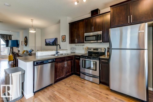 Kitchen featuring dark wood cabinetry, stainless steel appliances, light-toned countertops, wood-finish flooring, and a breakfast bar - 67 603 Watt Boulevard, Edmonton, AB - Indoor Photo Showing Kitchen