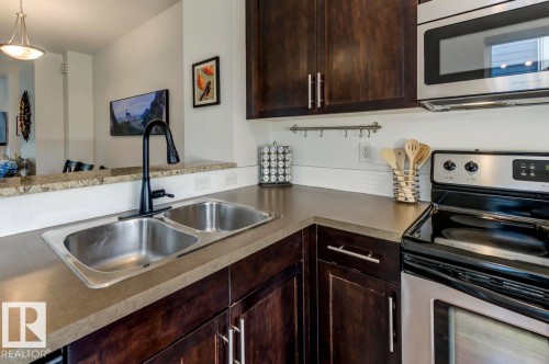 Modern kitchen featuring a double stainless steel sink with a matte black faucet, dark wood-finish cabinetry, light-toned countertops, a stainless steel microwave, and a stainless steel and black range - 67 603 Watt Boulevard, Edmonton, AB - Indoor Photo Showing Kitchen With Double Sink