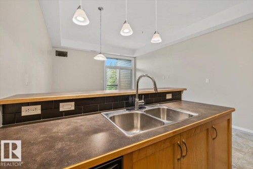 Kitchen island featuring a double basin stainless steel sink with a high-arc faucet, durable countertop, and wood-finish cabinetry - 228 10531 117 Street, Edmonton, AB - Indoor Photo Showing Kitchen With Double Sink