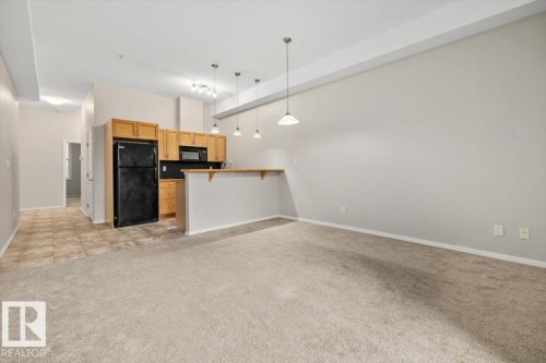Open-concept kitchen and living area featuring blonde wood cabinetry, black appliances, a breakfast bar, pendant lighting, and carpet flooring in the living space - 228 10531 117 Street, Edmonton, AB - Indoor Photo Showing Kitchen